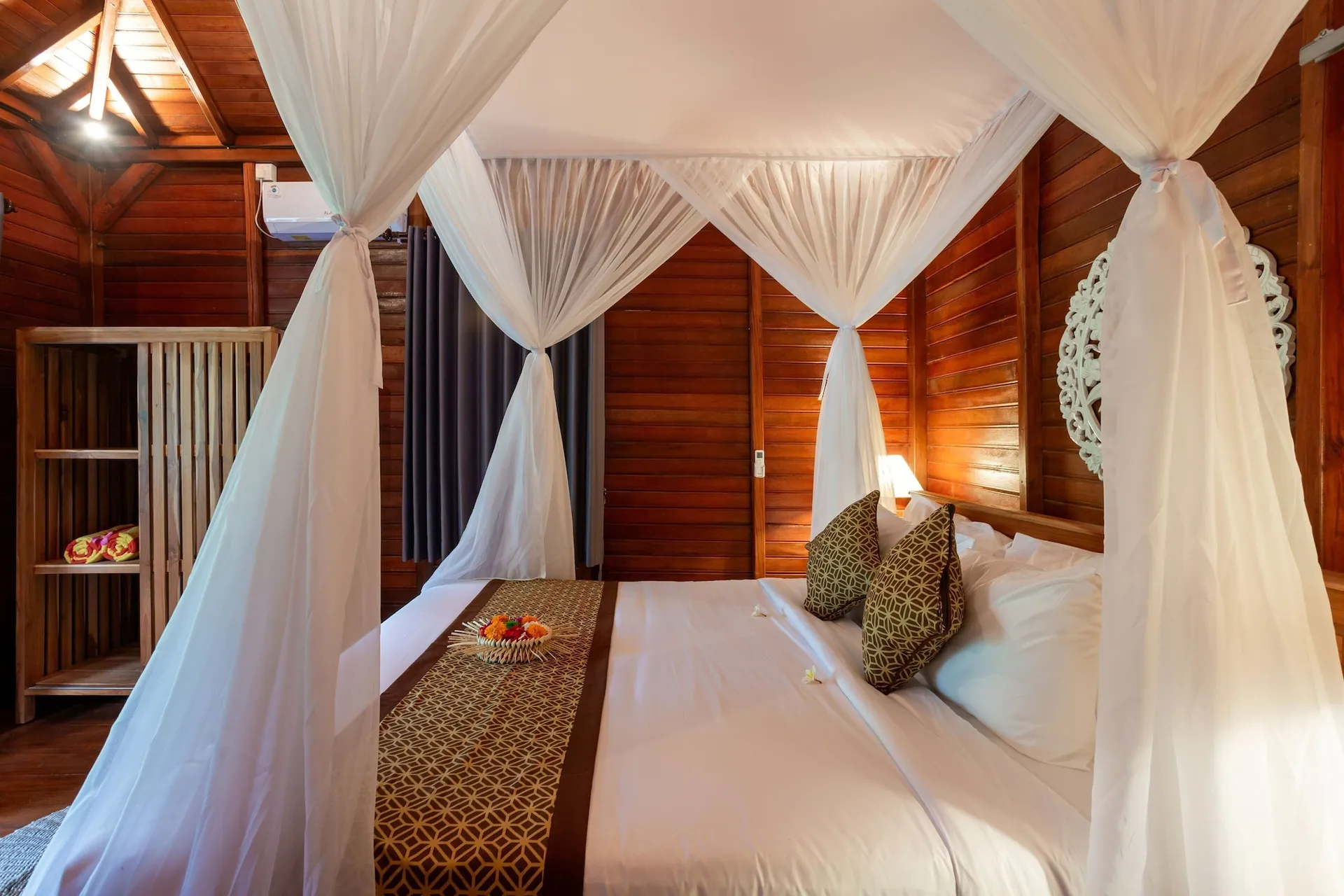 A cabin bedroom with a white-draped four-poster bed, timber walls and ceiling, and a carved wooden panel above the headboard