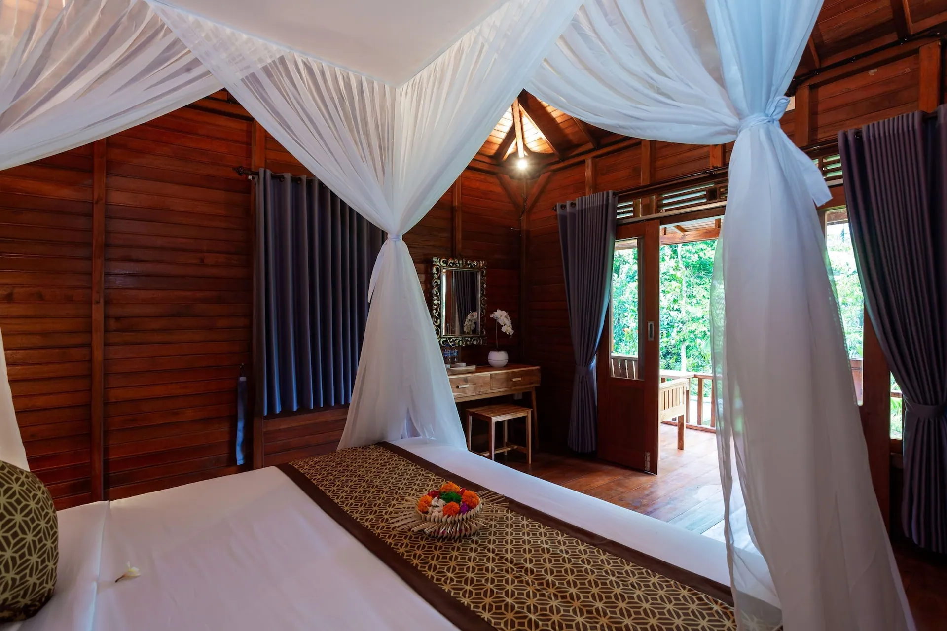A cabin interior with the bed beside a curtained window, timber walls throughout and a batik runner across the bed