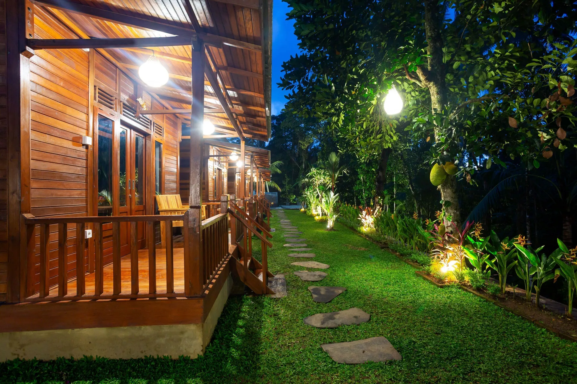A cabin's sheltered veranda lit warmly at dusk, with its timber balustrade and steps down to the lawn