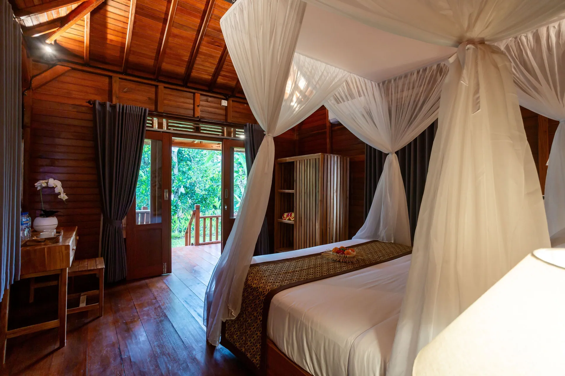 Inside a cabin looking towards the open door, the four-poster bed draped in white mosquito netting and green garden visible outside