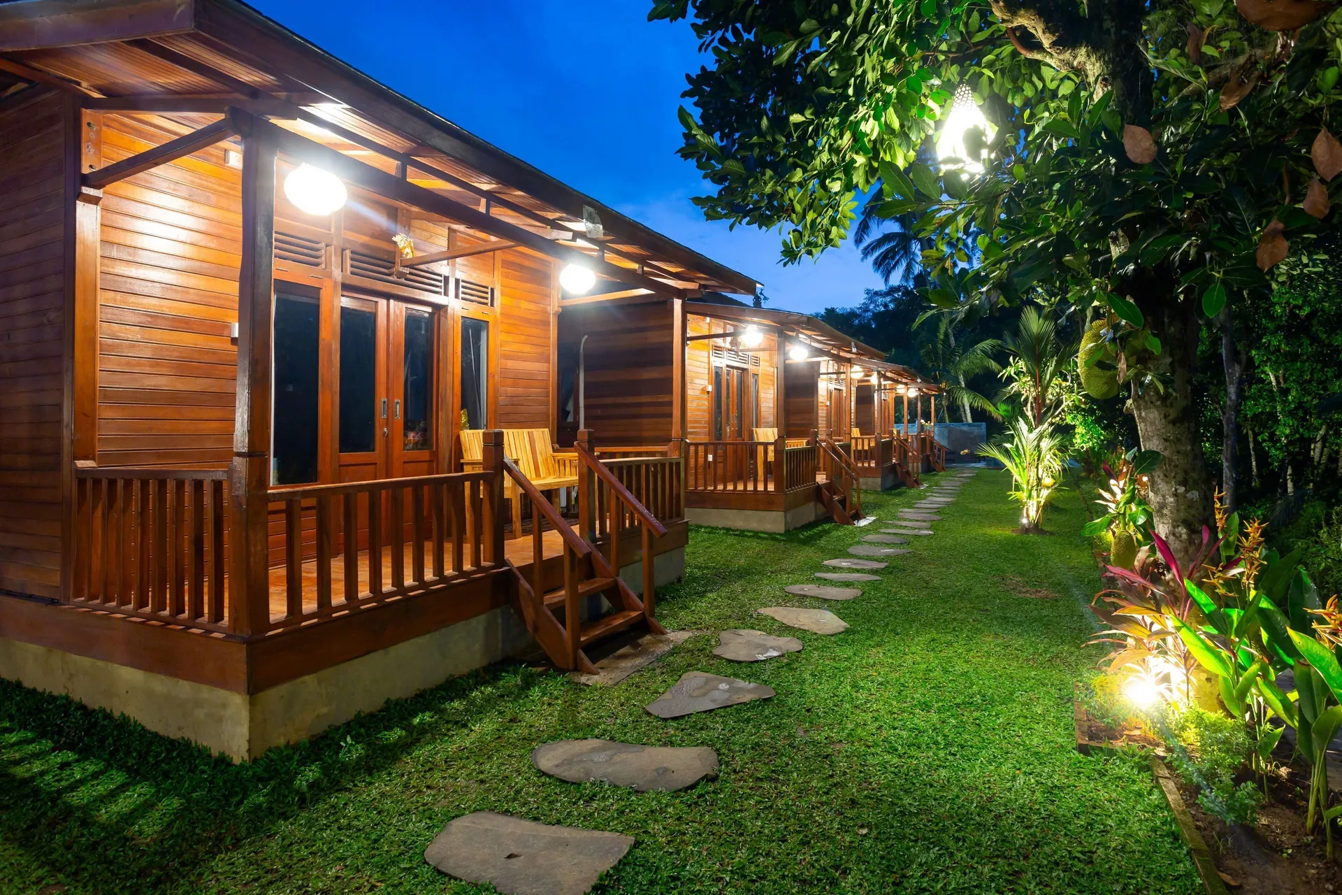 A stepping-stone path running between the lawn and the row of lit timber cabins in the evening, planting glowing along the edge